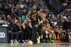 A basketball player in a black jersey with "Cavs" logo dribbles the ball during a game, with spectators and teammates in the background.