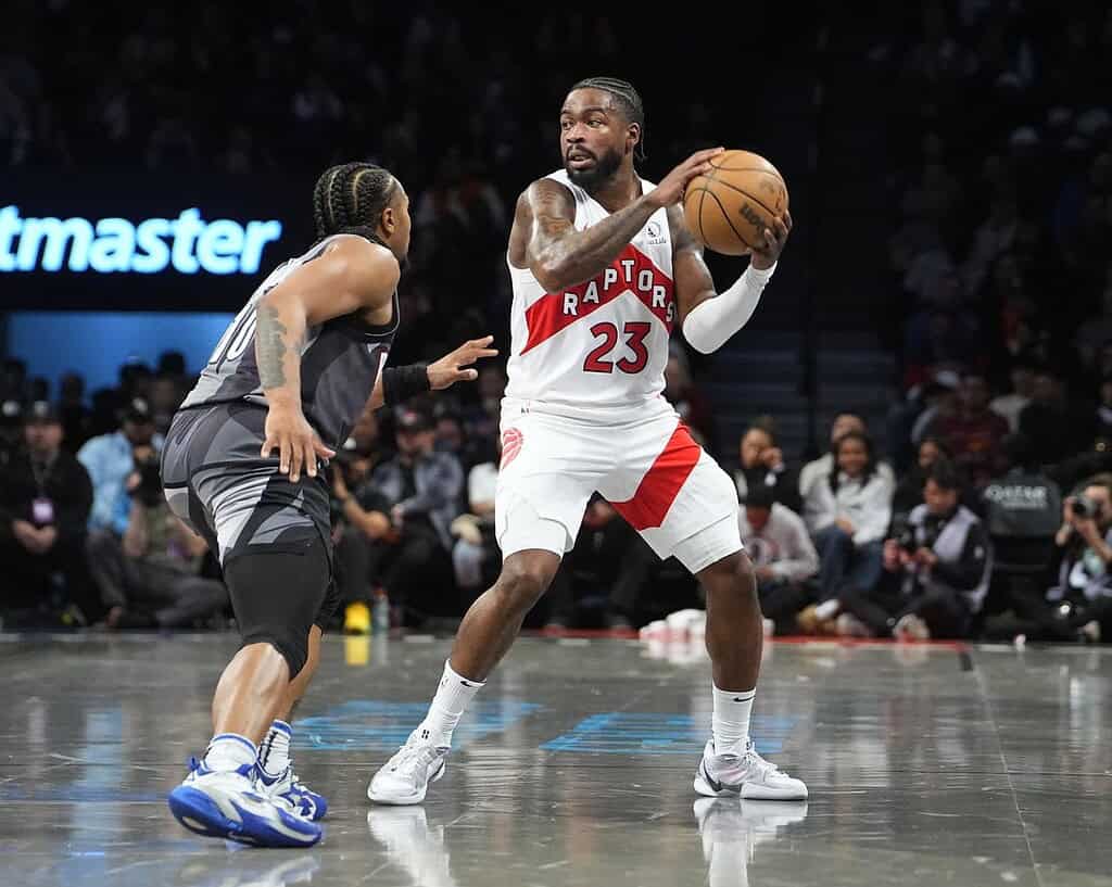 A Toronto Raptors player wearing white and red jersey prepares to pass while being closely guarded by an opponent in a basketball game.