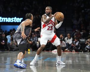 A Toronto Raptors player wearing white and red jersey prepares to pass while being closely guarded by an opponent in a basketball game.