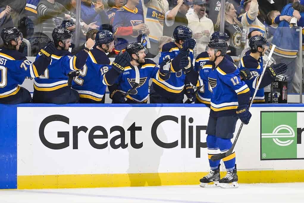 A group of hockey players in blue jerseys celebrate on the bench, with cheering fans visible in the background.