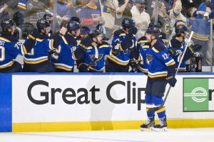 A group of hockey players in blue jerseys celebrate on the bench, with cheering fans visible in the background.
