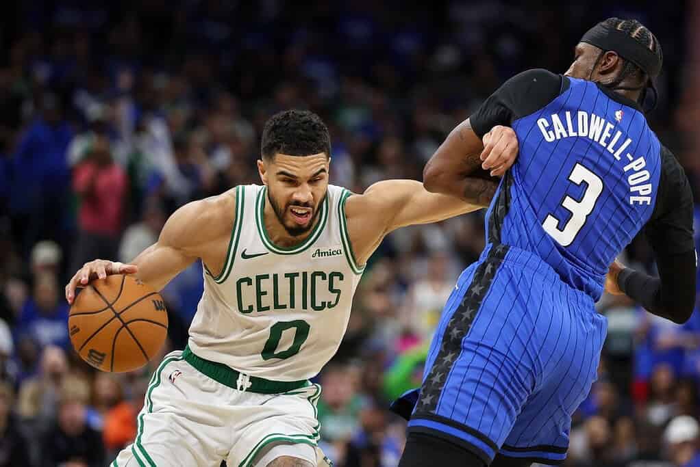 A Boston Celtics player dribbles the basketball while closely guarded by a player wearing a black and blue jersey with "CALDWELL-POPE" on the back.