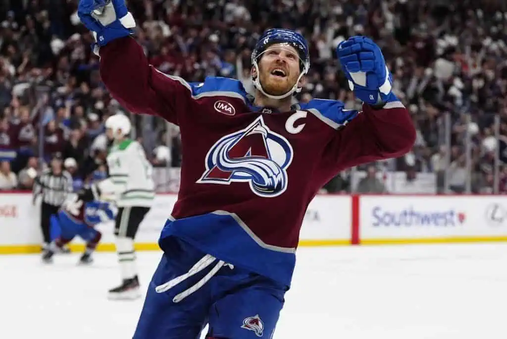 A hockey player in a maroon jersey celebrates a goal, raising his arms in triumph amidst a cheering crowd.
