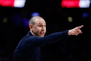 A coach passionately directs his team during a basketball game, emphasizing a point with an outstretched arm against a vibrant backdrop.