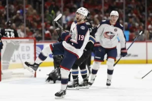 A hockey player celebrates a goal on the ice while teammates and opponents are in the background, amidst a packed arena.