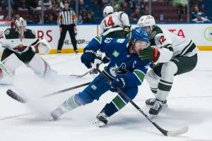 A player in a blue hockey jersey struggles for control of the puck amidst a flurry of ice, with opponents in green nearby.