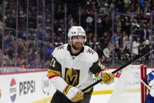 A hockey player wearing a white jersey with a gold logo celebrates a goal, surrounded by cheering fans in a lively arena.