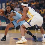 A basketball player in a blue jersey holds the ball, closely guarded by an opponent in a white jersey, during an intense game moment.