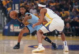 A basketball player in a blue jersey holds the ball, closely guarded by an opponent in a white jersey, during an intense game moment.