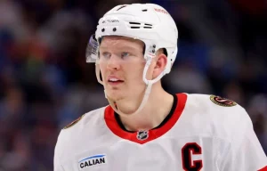 A hockey player in a white jersey with red accents, wearing a helmet and visor, stands on the rink, exuding focus and intensity.