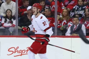A hockey player in a red and white jersey celebrates on the rink, surrounded by fans in team jerseys at a lively game.
