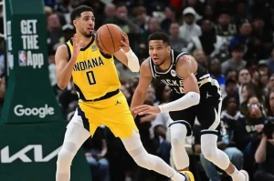 A player in a yellow Indiana uniform dribbles a basketball while a defender in a black and white jersey attempts to steal it.