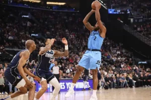 A basketball player in a light blue uniform takes a jump shot, while two defenders in dark uniforms attempt to block him.