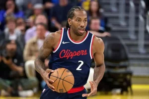A basketball player in a Clippers jersey dribbles the ball during a game, surrounded by seated fans in the background.