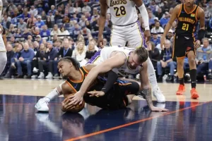 Two basketball players struggle for the ball on the court, surrounded by teammates and an engaged crowd in the background.