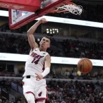 A basketball player in a white "Miami Heat" jersey performs a powerful dunk during a game, with fans visible in the background.