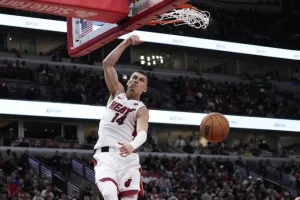 A basketball player in a white "Miami Heat" jersey performs a powerful dunk during a game, with fans visible in the background.