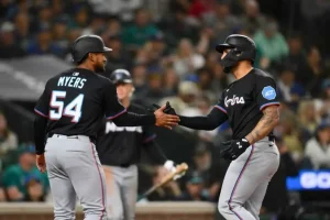 Two baseball players in black uniforms share a handshake and celebrate a play on the field, with fans visible in the background.