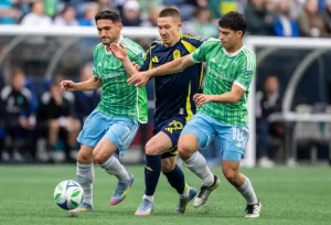 Two soccer players in green and blue jerseys are engaged in a competitive match, attempting to gain control of the ball from an opponent.