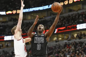 A basketball player in a black Nets jersey shoots a jump shot, while an opponent reaches to block the attempt in a packed arena.