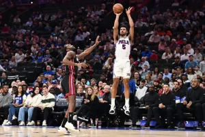A player in a white Philadelphia 76ers uniform jumps to take a shot during a basketball game, with spectators in the background.
