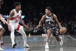 A player in a black Brooklyn jersey dribbles a basketball, while two defenders in white Raptors jerseys closely guard him on the court.