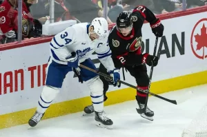 Two hockey players battle for the puck along the boards during a game, with one wearing a blue jersey and the other in black.