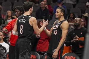 Two basketball players in black jerseys shake hands on the court, surrounded by teammates and fans in the stands.