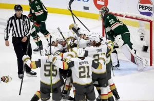 Hockey players celebrate a goal, surrounded by teammates, with an official nearby and a goaltender watching from the crease.