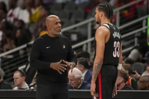 A basketball coach and player engage in discussion on the court, with fans watching in the background. The player wears a team uniform.