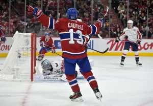 A Montreal Canadiens player, Caufield, celebrates a goal as the puck is in front of the goalie, amidst a packed arena.