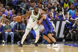 A basketball player in a Celtics jersey dribbles the ball while being closely guarded by an opponent in a blue uniform during a game.