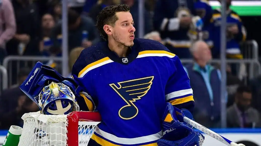 Winnipeg Jets vs St. Louis Blues Picks and Predictions May 4th 2025 2 A hockey goalie in a St. Louis Blues jersey leans against the goal post, holding a helmet, with spectators in the background.