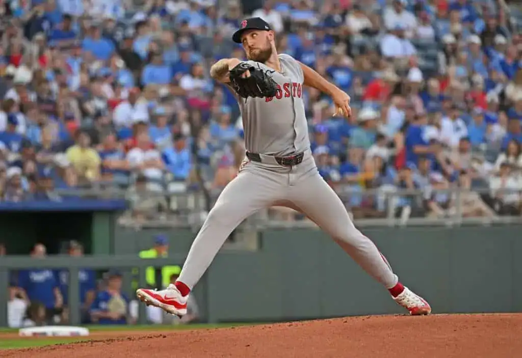 A Boston Red Sox pitcher in mid-throw on a baseball mound, with a crowd of fans in the background at a sunny game.