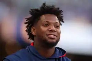 A person with curly hair wearing a navy hoodie with red accents, set against a blurred background of a sports event.
