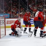 A tense moment in a hockey game, with players in red jerseys battling near the goal while the goalie is ready in the net.