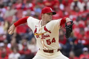 A St. Louis Cardinals pitcher in a white uniform with red accents is preparing to throw a baseball on the field, surrounded by a cheering crowd.
