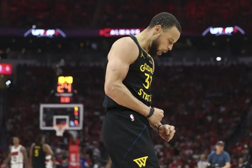 A basketball player in a Golden State Warriors jersey celebrates a successful play during an intense game, fans cheering in the background.