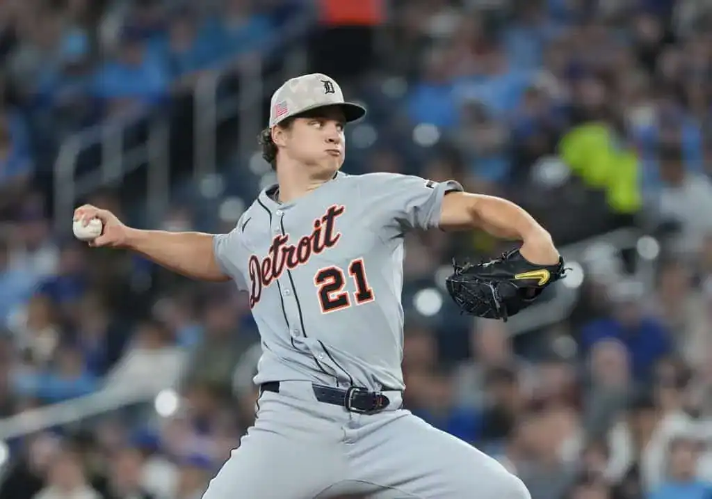 A baseball player in a uniform throws a ball on an outdoor baseball field, surrounded by stadium seating and baseball equipment.