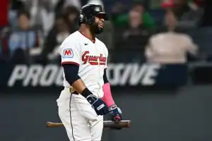 A baseball player wearing a Guardians jersey stands on the field, holding a bat and wearing pink batting gloves, ready to play.