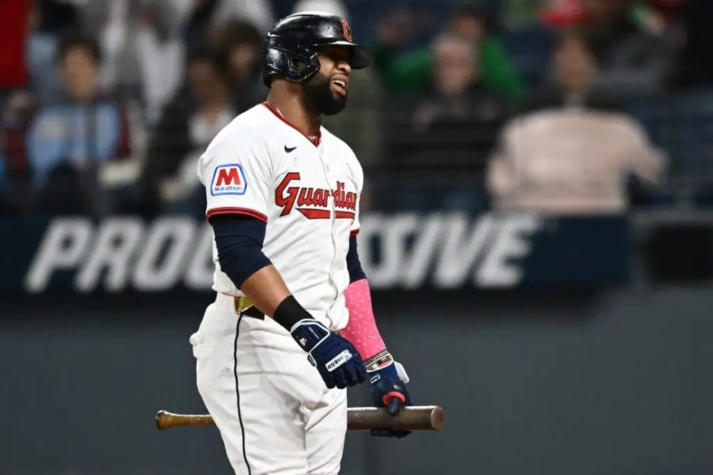 A baseball player wearing a Guardians jersey stands on the field, holding a bat and wearing pink batting gloves, ready to play.