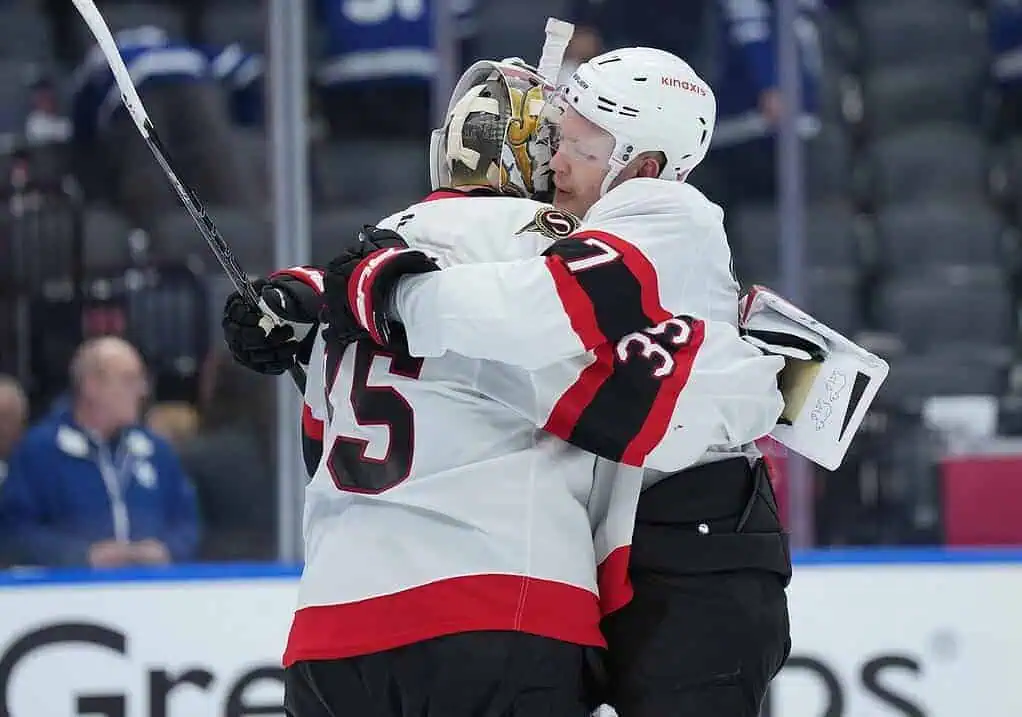 Two hockey players embrace in celebration, wearing red and white jerseys, with one holding a clipboard, in a stadium setting.