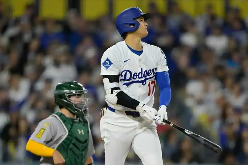 A Los Angeles Dodgers player in a white jersey and blue cap swings a bat, while a green-uniformed catcher looks on during a game.