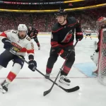 Two hockey players compete for the puck on the ice, while a goalie is positioned near the net in the background amid a vibrant crowd.