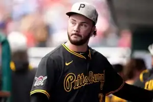 A baseball player in a black Pittsburgh Pirates jersey and cap stands in a dugout area, surrounded by blurred teammates and fans.