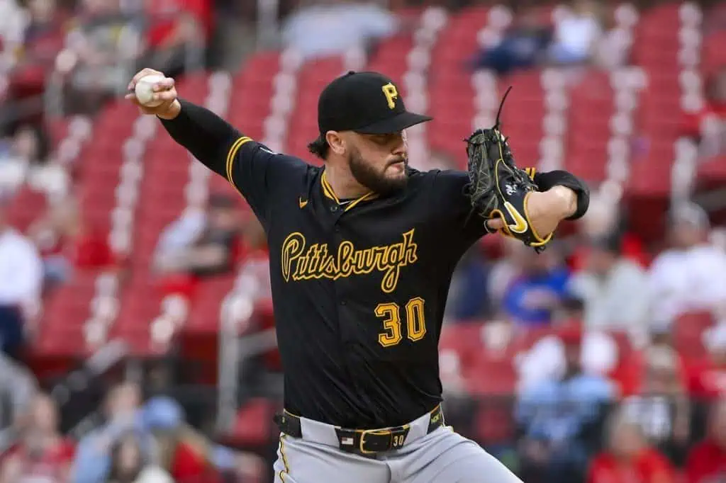 A pitcher in a black Pittsburgh Pirates jersey winds up to throw a baseball in a ballpark with empty red seating in the background.