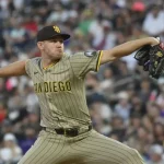 A pitcher in a tan San Diego Padres uniform winds up to throw a baseball, with a blurred crowd of fans cheering in the background.