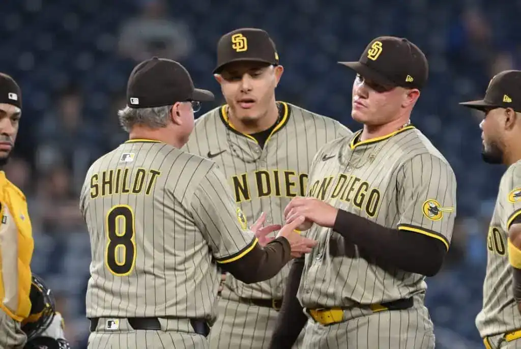 Baseball team huddled on the field, discussing strategy. Players wear gray uniforms with black and yellow accents.