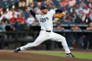 A pitcher in a white "Rays" jersey prepares to throw a baseball on the mound, with a crowd of fans visible in the background.
