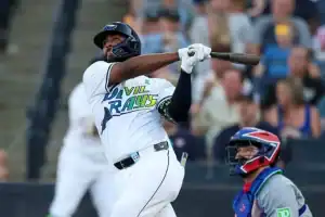 A baseball player in a blue and white jersey swings a bat during a game, with an audience visible in the background.